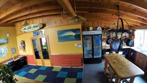 A cozy room with wooden beams, a wooden table, a bench, a refrigerator, and a wall with a painting and a sign. - Bazil's Hostel & Surf School (Westport)