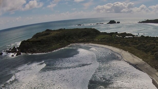 An aerial view of a coastal area with a road curving along the beach, rocky outcrops, and a clear view of the ocean. - Bazil's Hostel & Surf School (Westport)