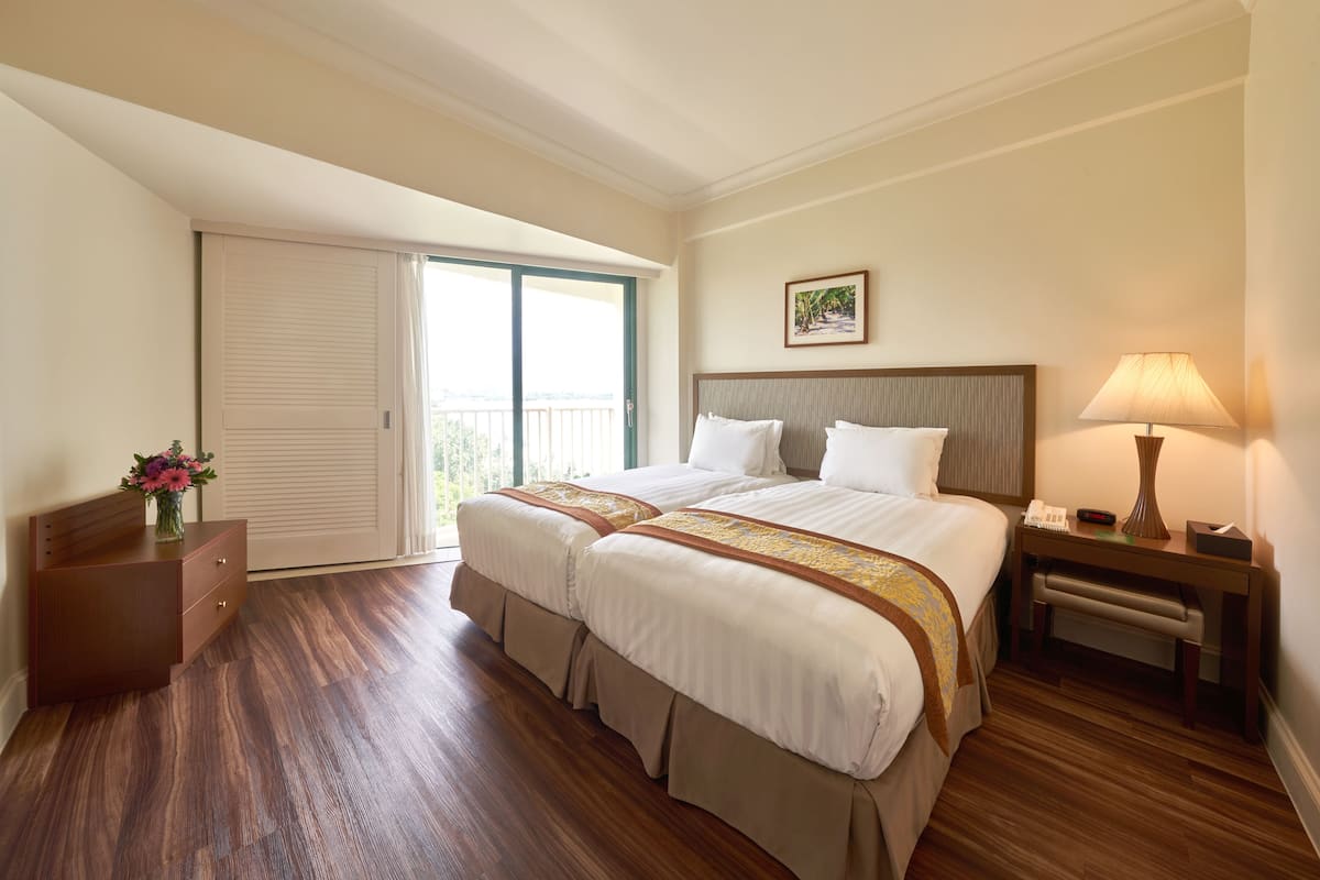 A hotel room with two beds, a nightstand, a lamp, and a view of the outdoors.