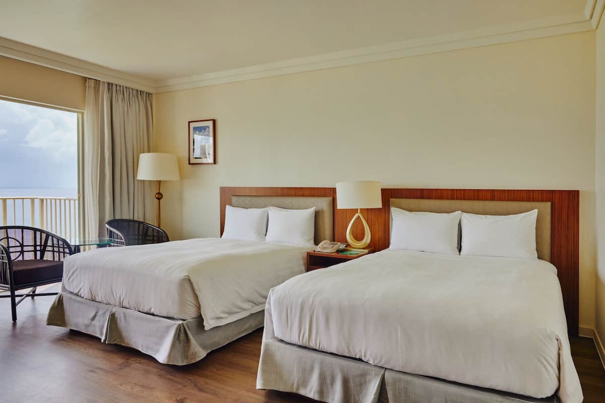 A hotel room with two beds, a chair, a nightstand with a lamp, and a view of the sea.