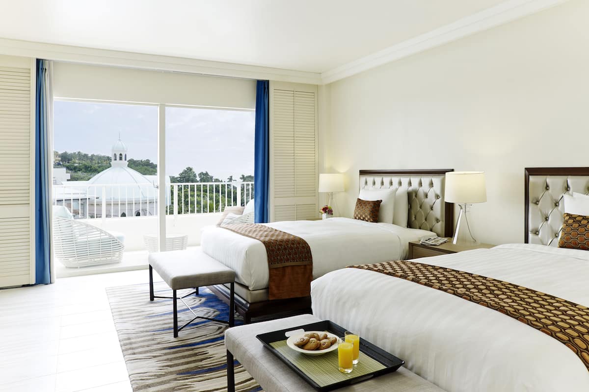 A hotel room with two beds, a large window with a view of a white building, and a tray with cookies and juice on the bed.