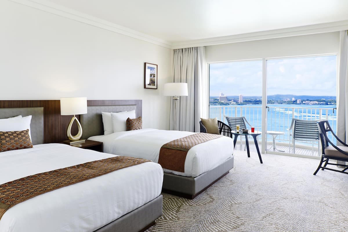 A hotel room with two beds, a balcony with a view of the city and water, and a small table with chairs.