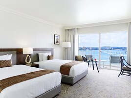 Habitación de hotel con dos camas, un balcón con vistas a la ciudad y al agua, y una pequeña mesa con sillas.