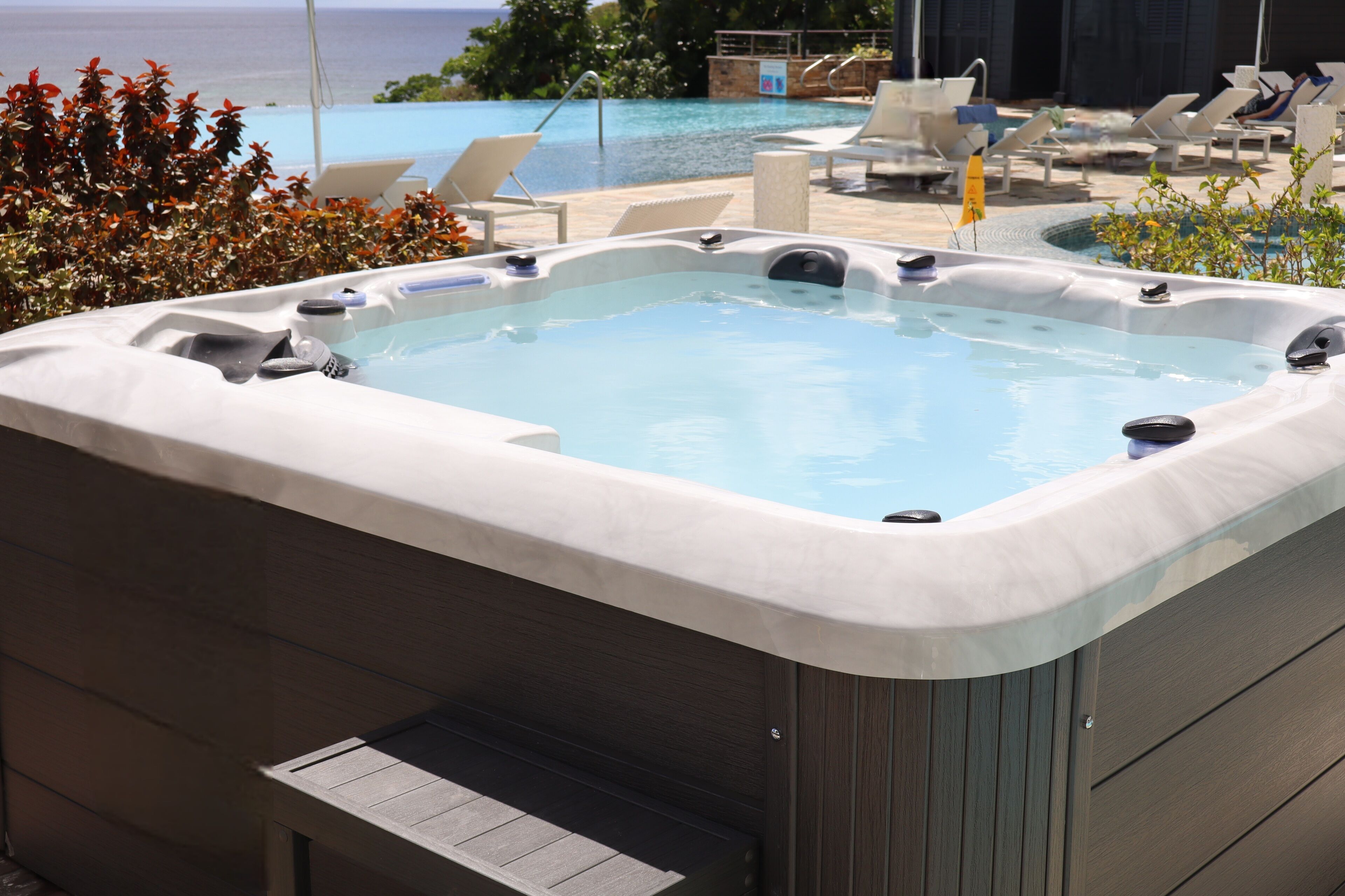 A hot tub with a view of the ocean, surrounded by lounge chairs and a pool.