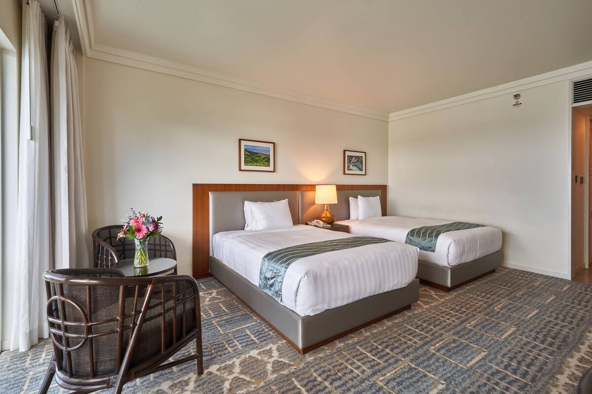 A hotel room with two beds, a small table with a vase of flowers, a chair, and a carpeted floor.