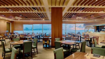 A spacious dining area with multiple tables and chairs, large windows offering a view of the sea, and a modern ceiling design.