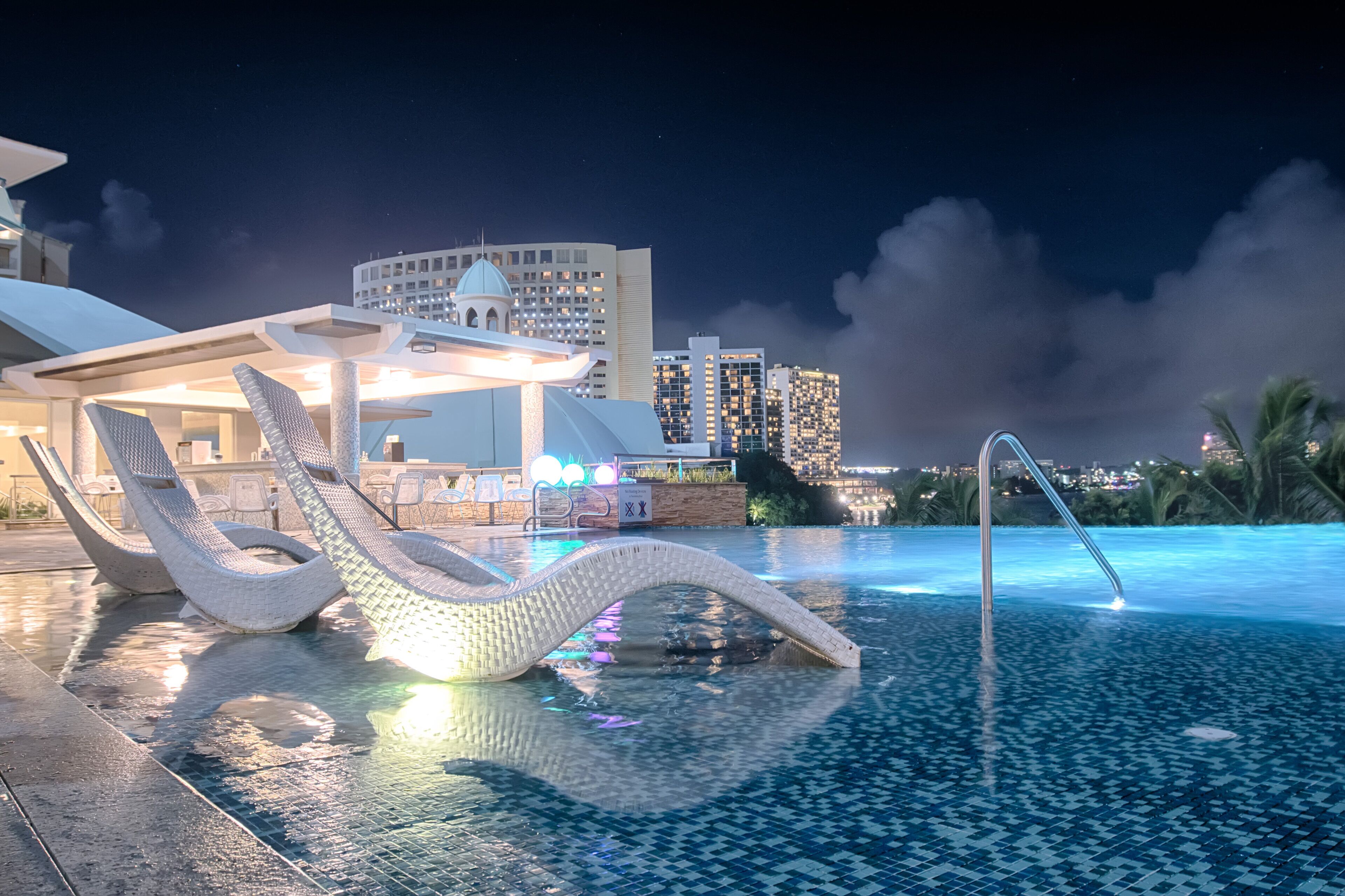 A nighttime pool area with lounge chairs and a view of a city skyline.