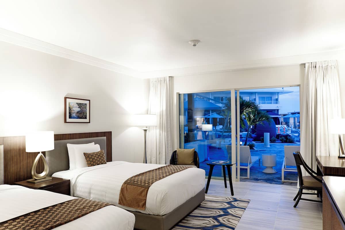A hotel room with two beds, a desk, a chair, a lamp, and a view of the pool area.