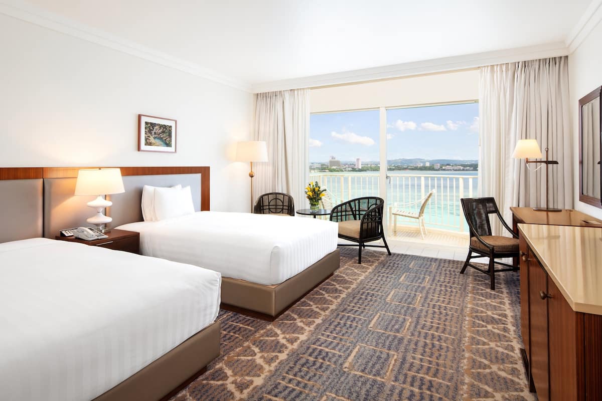 A hotel room with two beds, a desk with a TV, a chair, and a balcony with a view of the sea.