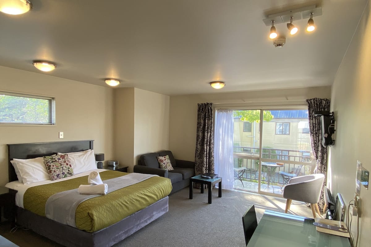 Une chambre d’hôtel avec un lit, un canapé, une petite table et une chaise. Il y a un balcon avec une table et des chaises à l’extérieur.