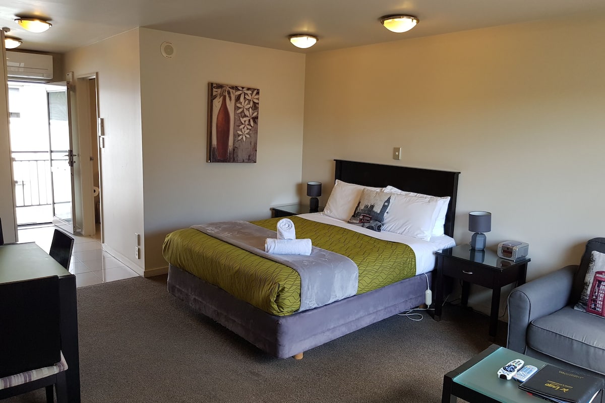 Une chambre d’hôtel comprenant un lit, un bureau, une chaise, un canapé et une petite table avec une télécommande et un livre.