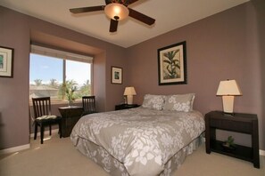 A bedroom with a large bed, two bedside tables with lamps, a ceiling fan, and a window with a view of palm trees.