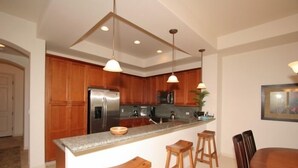 A modern kitchen with wooden cabinets, a stainless steel refrigerator, a granite countertop, and pendant lighting. - Fairways at Mauna Lani (Kamuela)