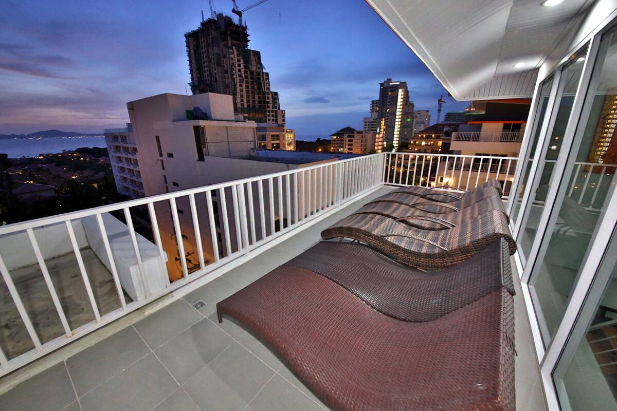 A balcony with a white railing, a brown woven sofa, and a view of the cityscape and water at dusk.