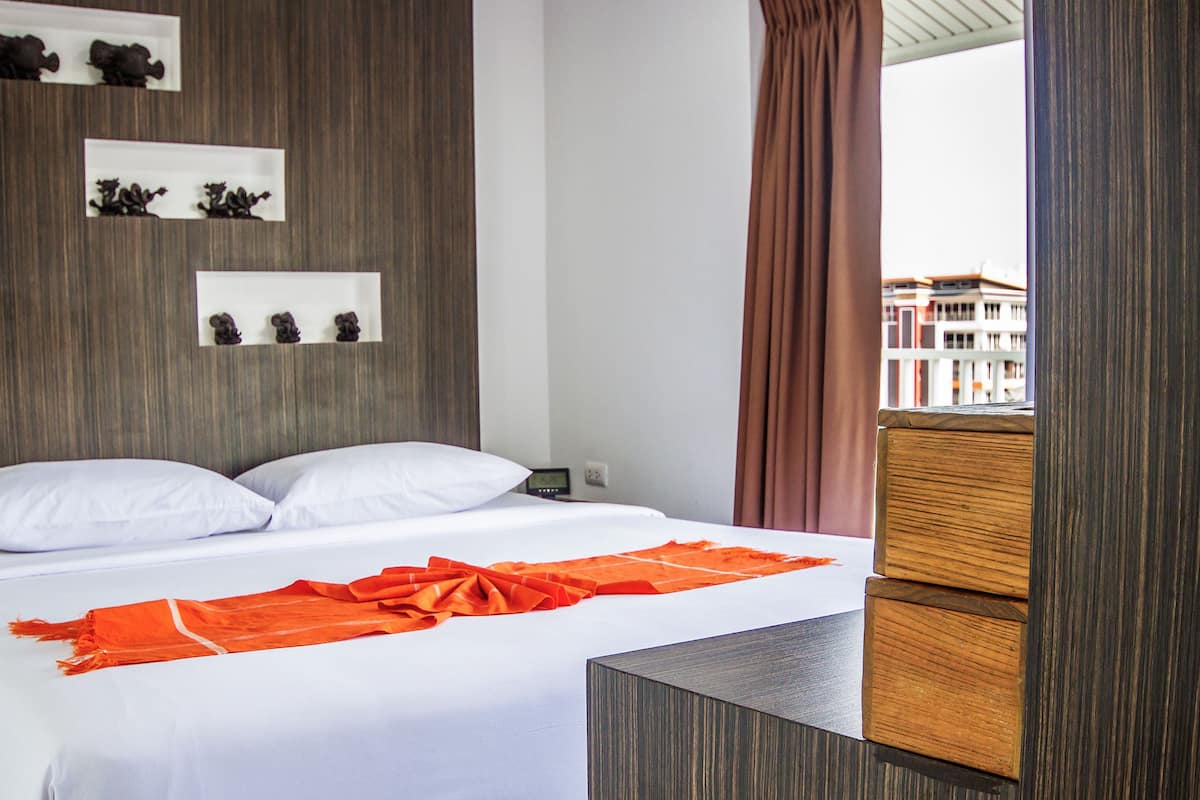 A hotel room with a bed, a wooden headboard, a nightstand, and a view of buildings through a window.