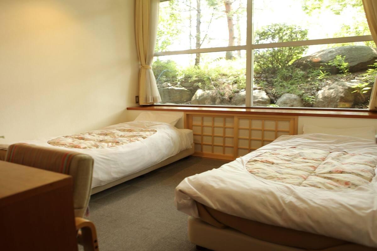 A room with two beds, a small table, and a window with a view of greenery.