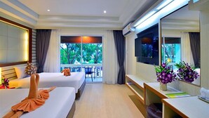 A hotel room with two beds, a TV, a desk, and a large window with a view of greenery. - Windmill Resort Hotel (Pattaya)