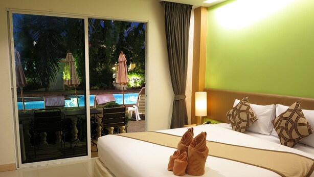 A hotel room with a bed, a nightstand, a lamp, and a view of an outdoor pool area. - Windmill Resort Hotel (Pattaya)