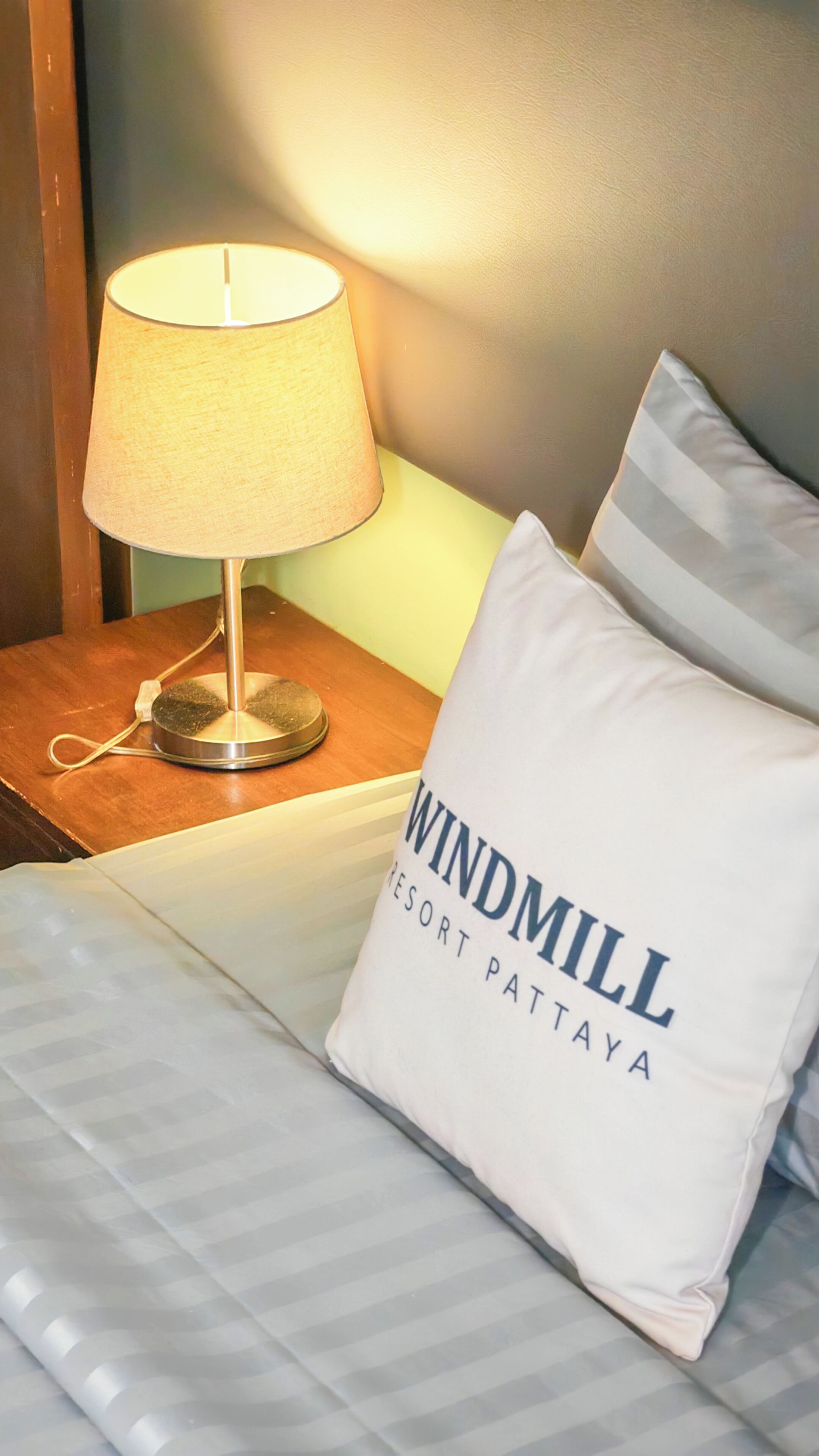 Ett hotellrum med en säng, en lampa och en kudde med texten ”Windmill Resort Pattaya”.