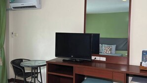 A hotel room with a television on a wooden desk, a small table with chairs, a green curtain, and a white air conditioning unit. - Windmill Resort Hotel (Pattaya)