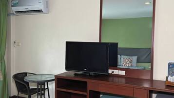 Habitación de hotel con una televisión sobre un escritorio de madera, una pequeña mesa con sillas, una cortina verde y un equipo de aire acondicionado blanco.