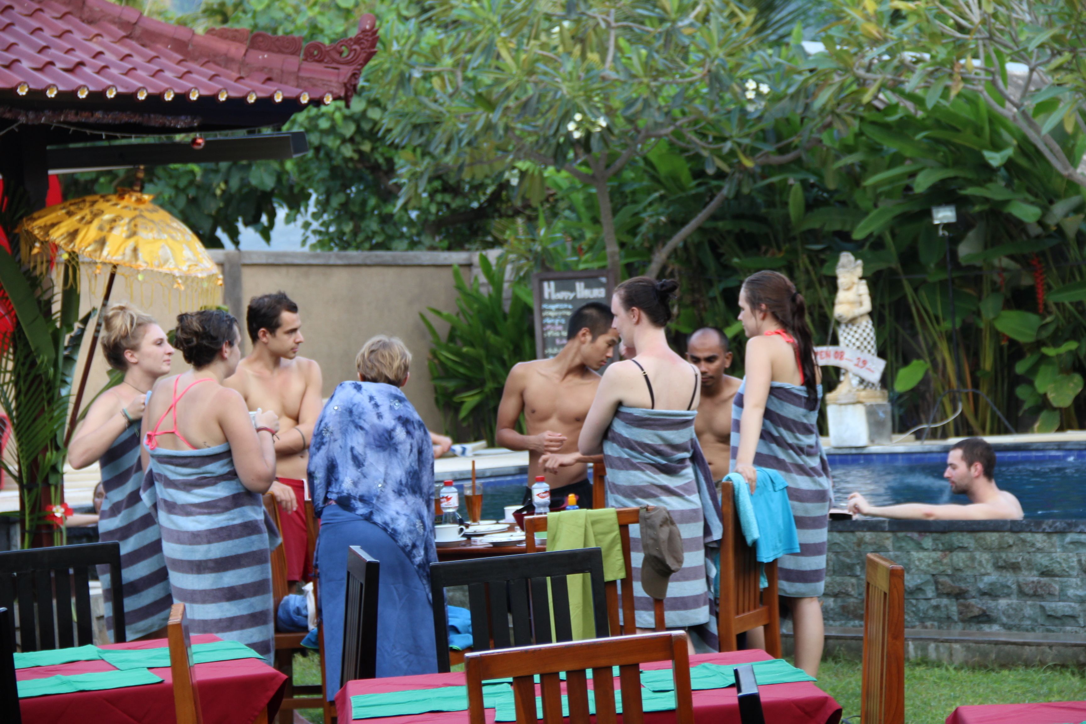 A group of people gathered around a poolside area with tables and chairs, some with umbrellas.