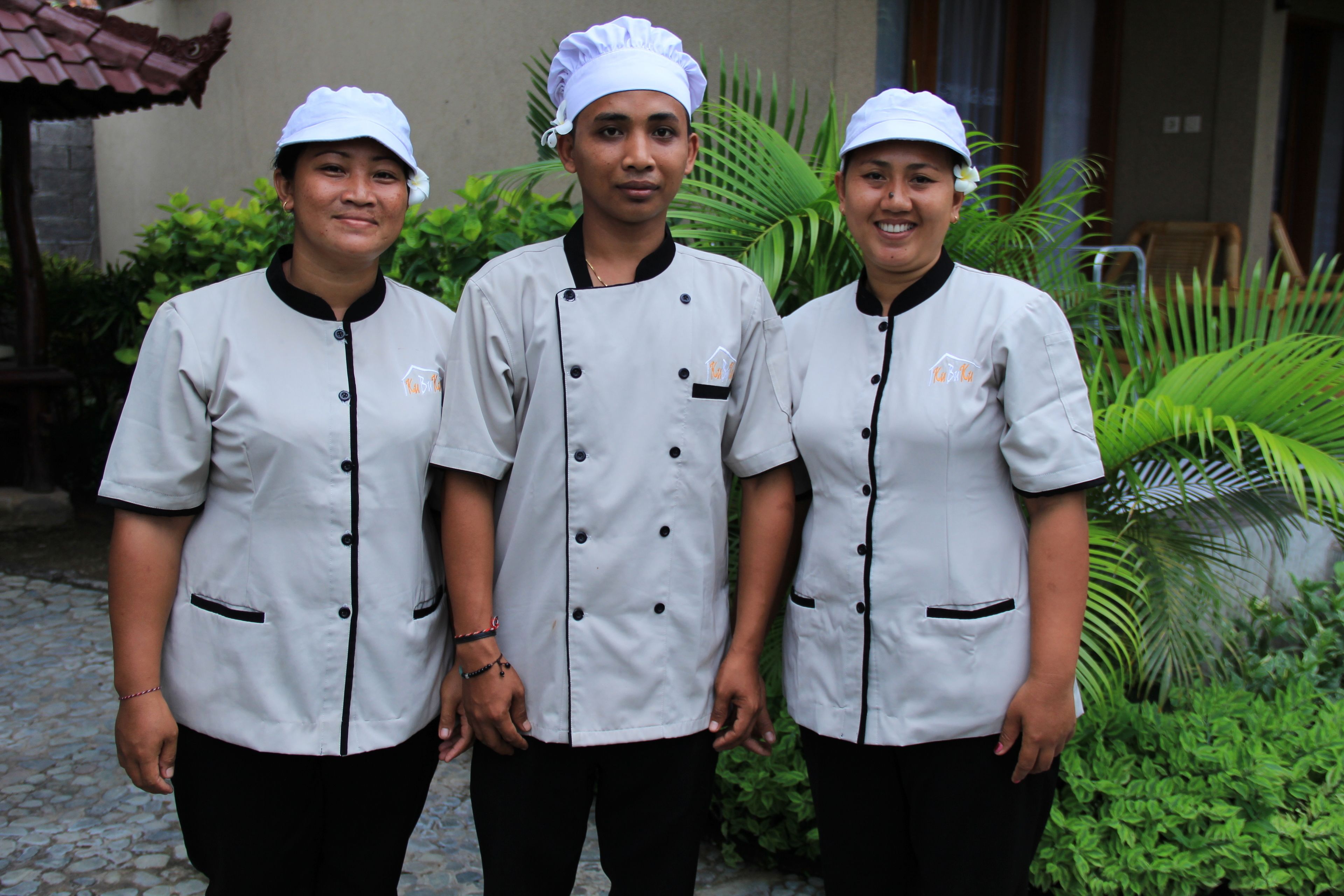 Three individuals in chef uniforms standing outdoors with greenery and a building in the background.