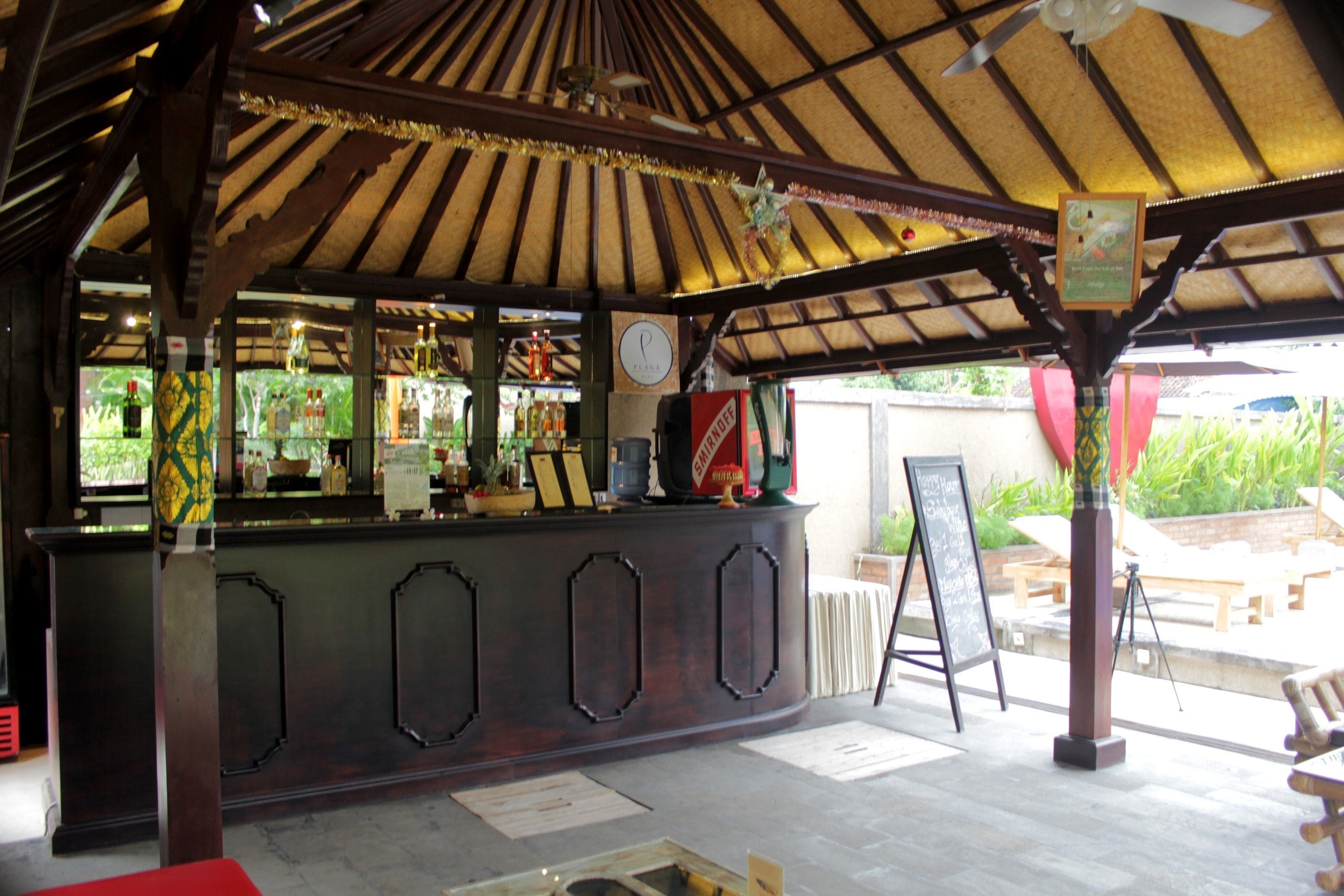 A covered outdoor bar area with a wooden ceiling and a counter displaying various bottles and a clock.