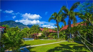 A lush garden with a swimming pool, palm trees, and a view of mountains under a clear sky.