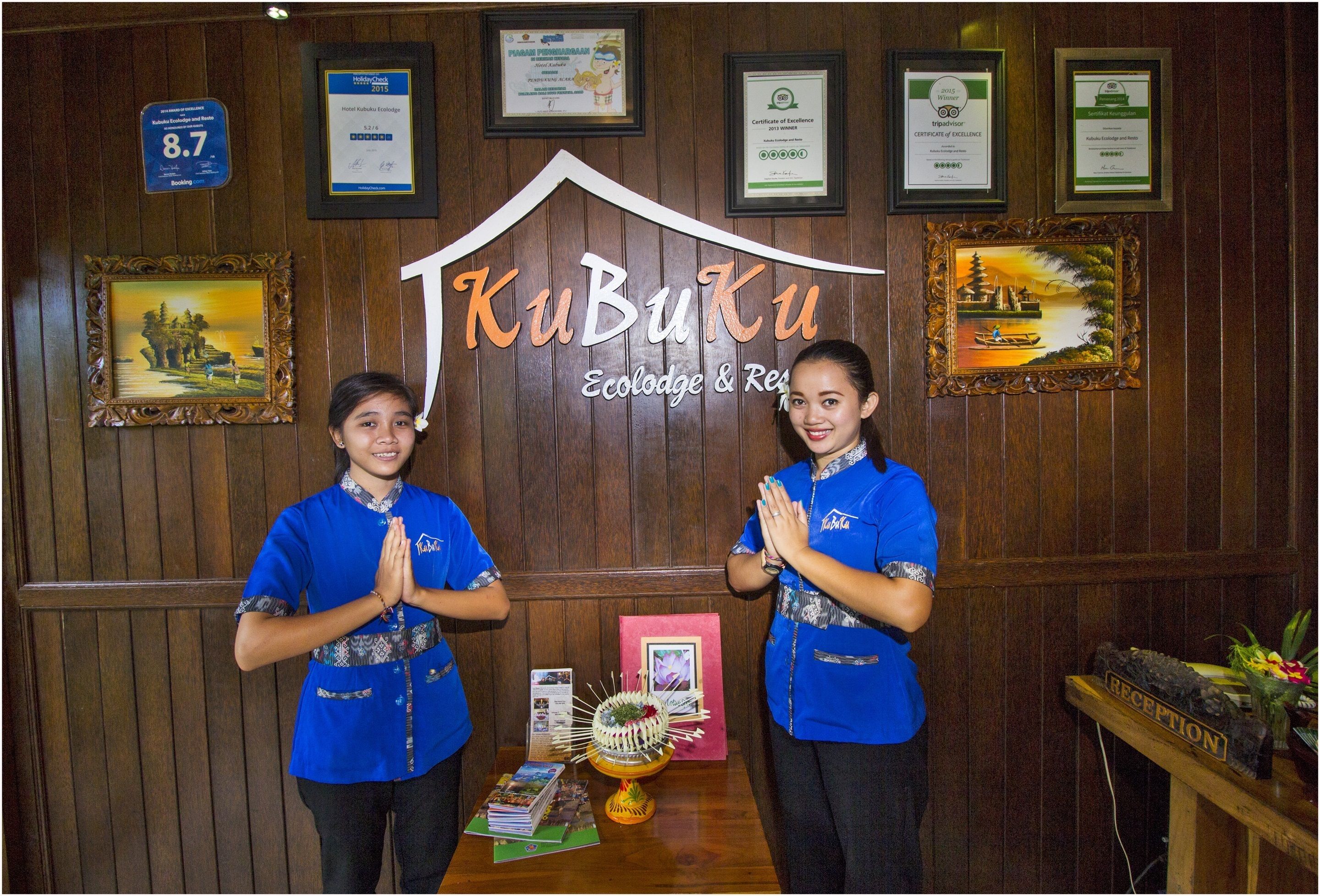 Dos personas con uniformes azules de pie frente a una recepción con un fondo de madera, mostrando varios certificados enmarcados y un letrero que dice "KuBuku Ecolodge & Resort".