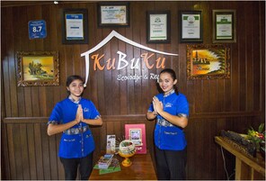 Dos personas con uniformes azules de pie frente a una recepción con un fondo de madera, mostrando varios certificados enmarcados y un letrero que dice "KuBuku Ecolodge & Resort".