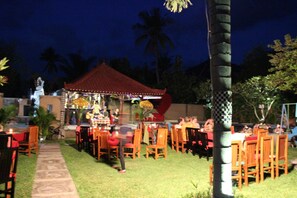 An outdoor dining area with tables and chairs, a covered seating area, and palm trees in the background. - Kubuku Eco Dive Lodge & Yoga (Pemuteran)