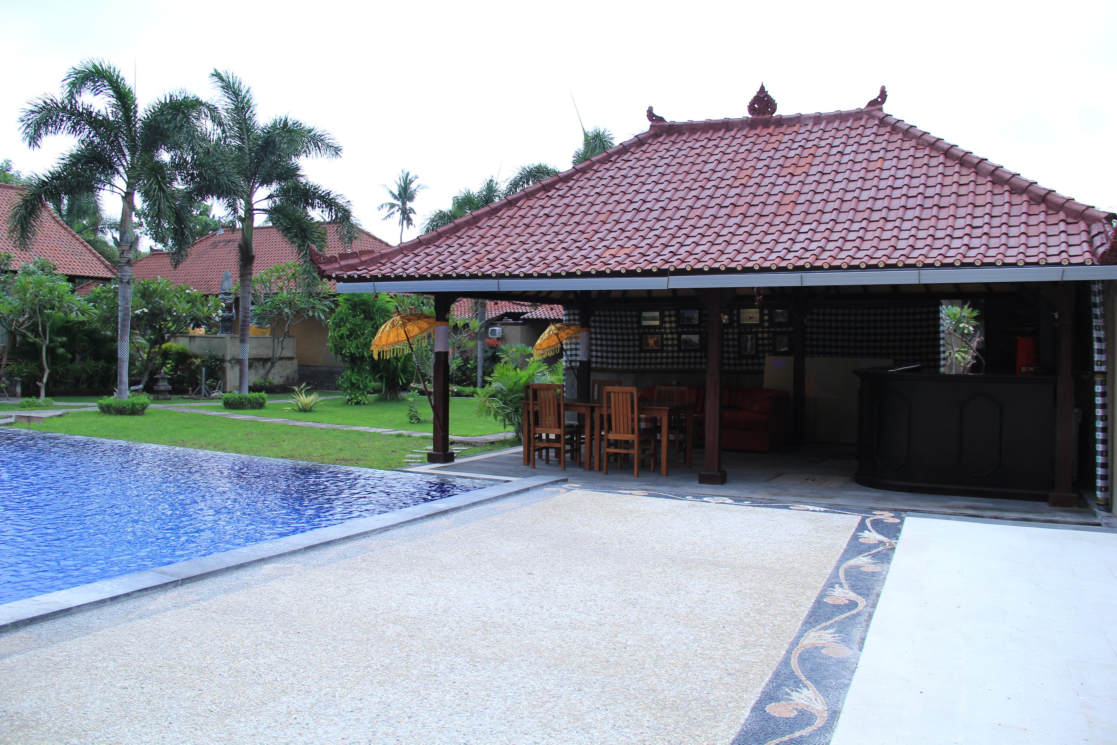 A pool area with a tiled roof structure, outdoor seating, and palm trees.