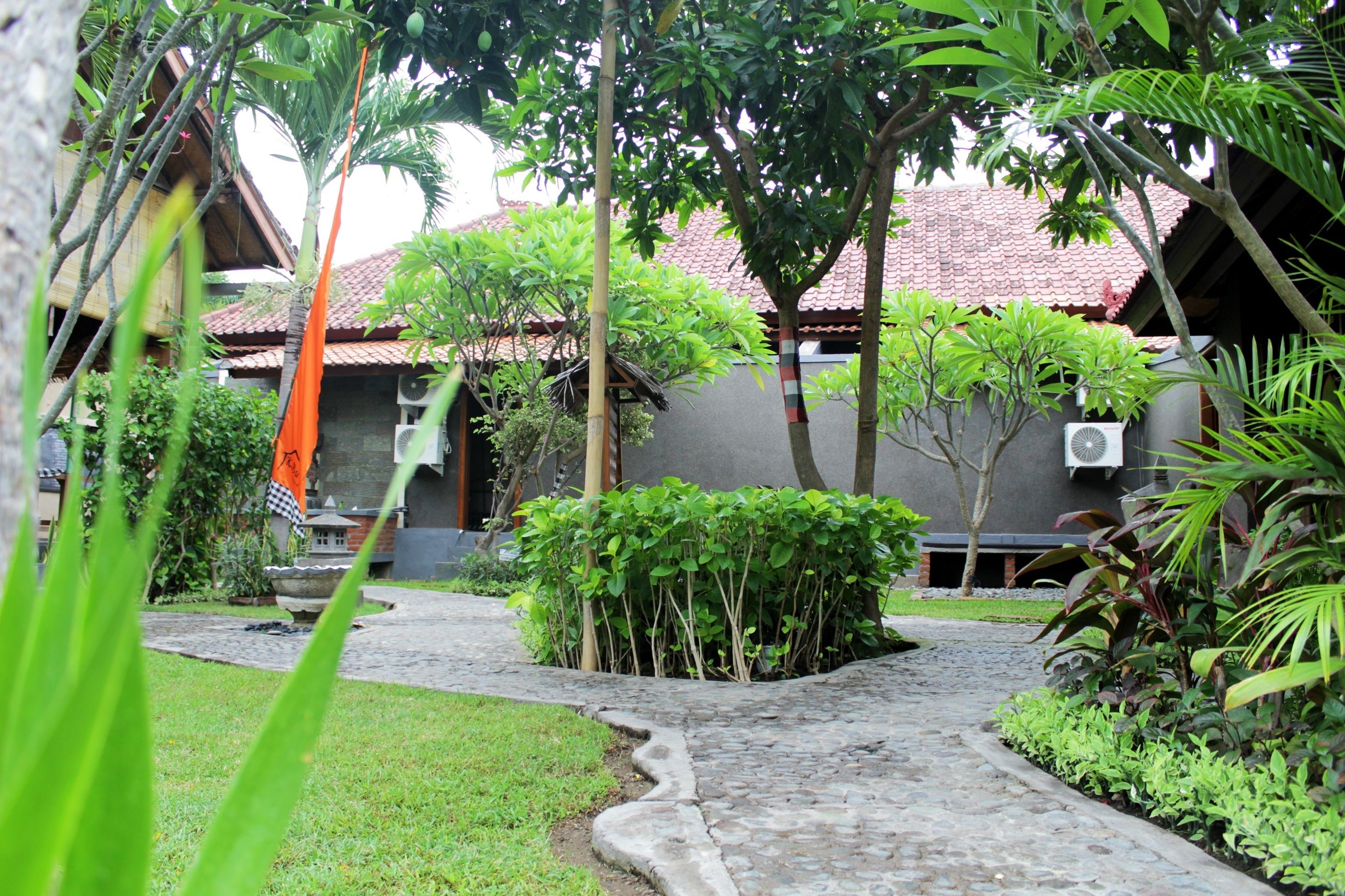 A courtyard with a cobblestone path, surrounded by lush greenery and a traditional tiled roof structure.