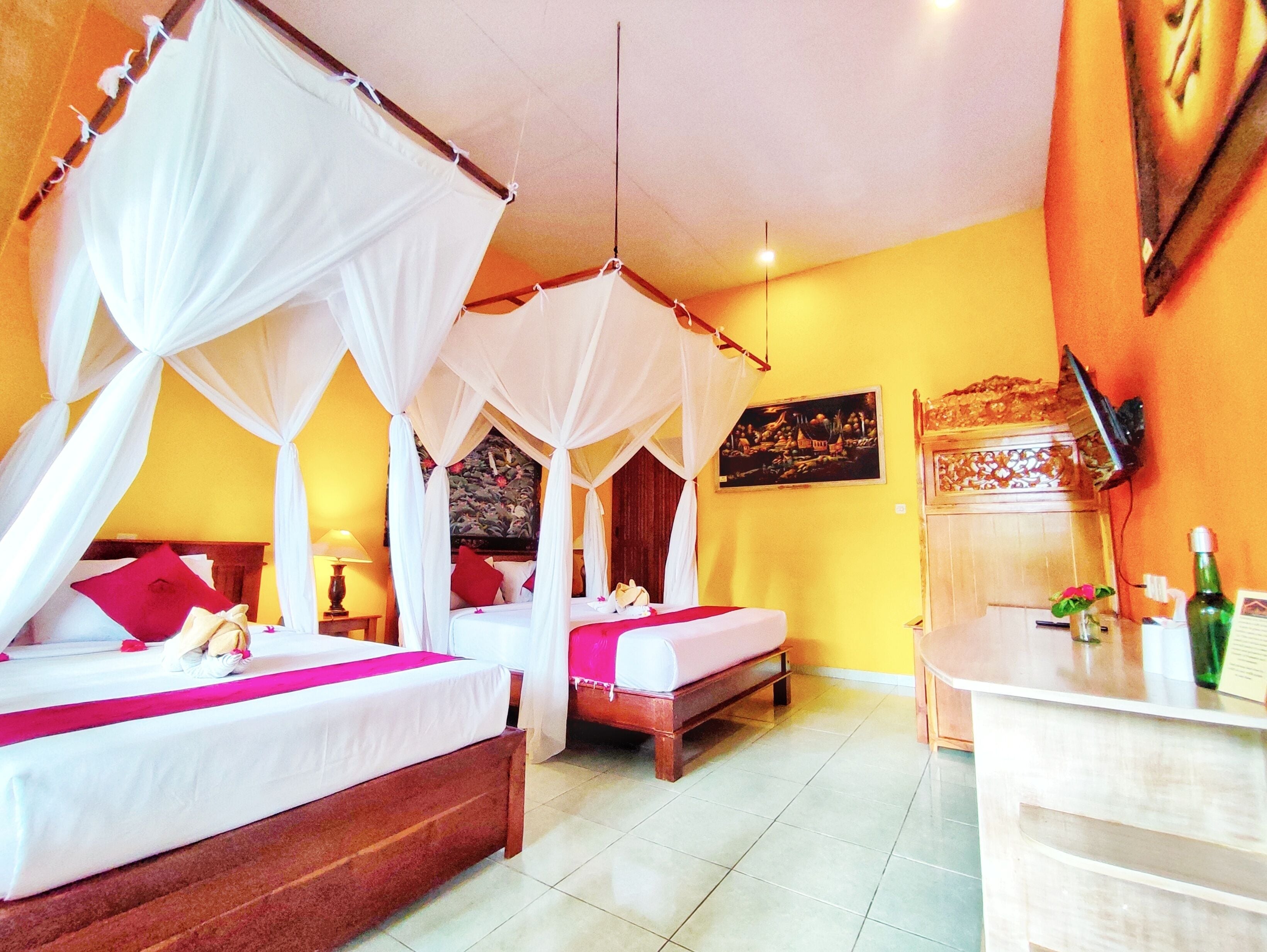 A hotel room with two beds, each with a canopy, a wooden headboard, and a red pillow. There is a small wooden table with a green bottle and a white bottle on it.