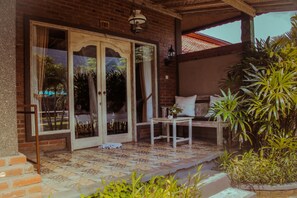 A covered patio with a tiled floor, a wooden bench, a small table with a plant, and a potted plant to the right. - Kubuku Eco Dive Lodge & Yoga (Pemuteran)
