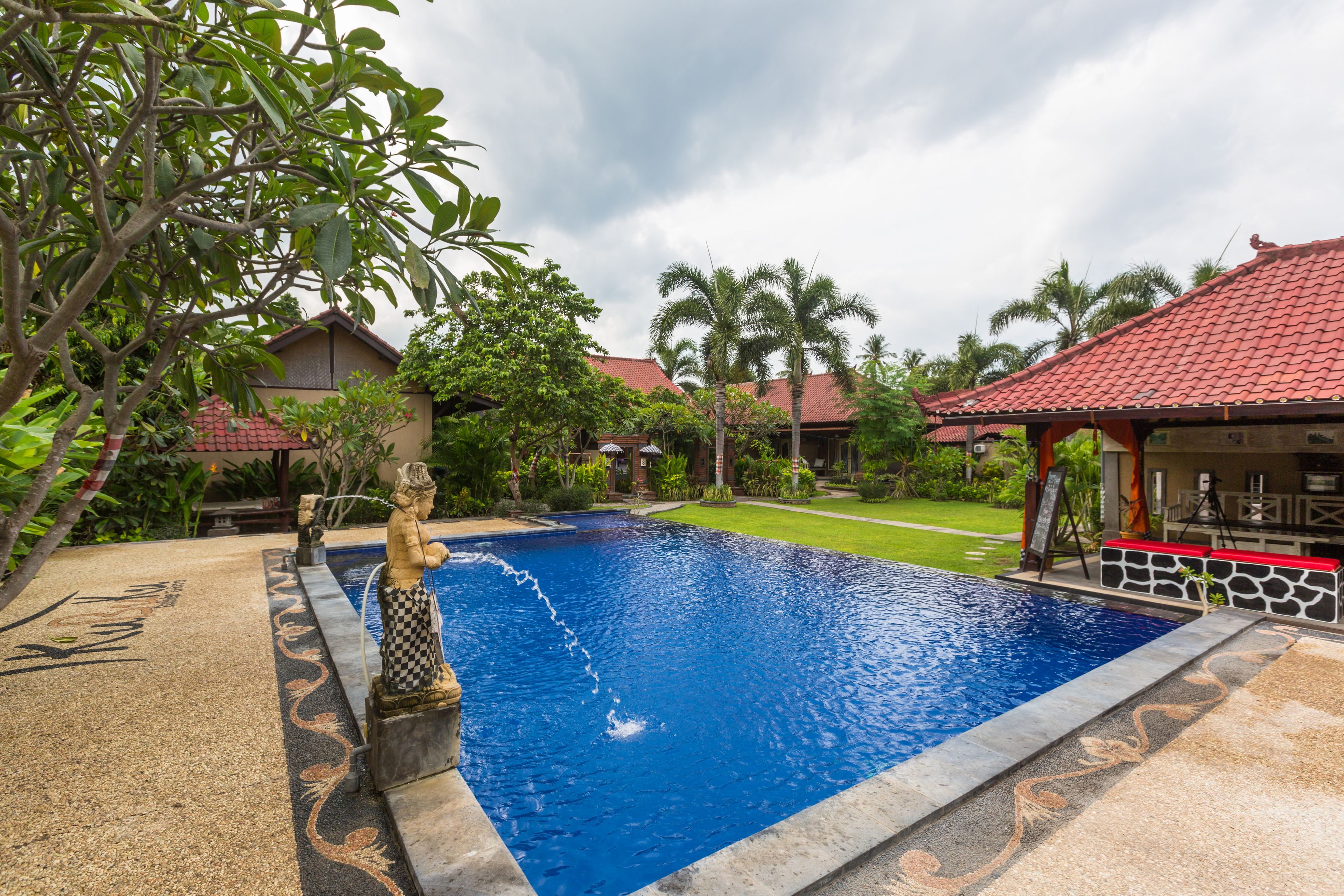 A swimming pool with a statue, surrounded by greenery and a red-roofed building.