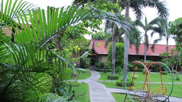 Stroll through the tranquil garden, featuring a traditional building with a red-tiled roof, lush tropical plants, and a children's swing set.