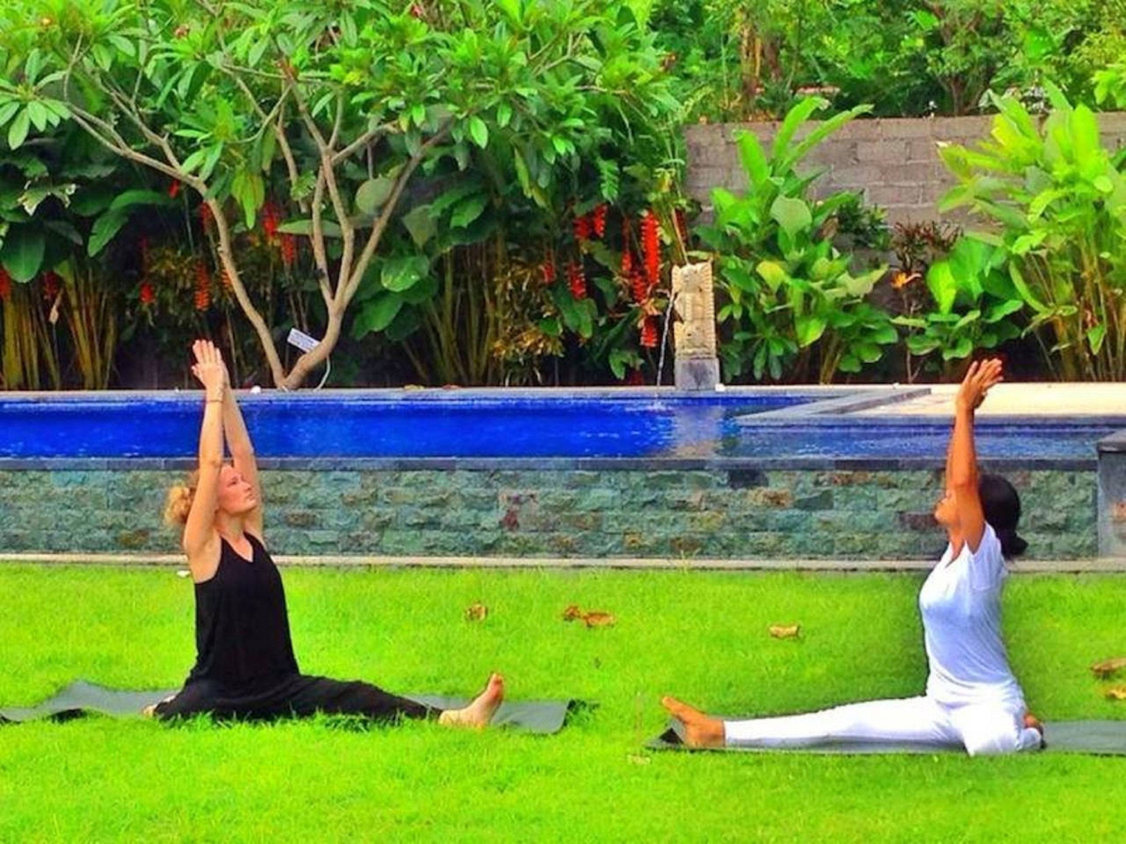 Deux personnes pratiquent le yoga sur l’herbe, avec un mur de pierre et de la verdure en arrière-plan.