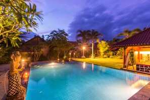 A swimming pool with a statue, a covered area, and a well-lit garden at dusk. - Kubuku Eco Dive Lodge & Yoga (Pemuteran)
