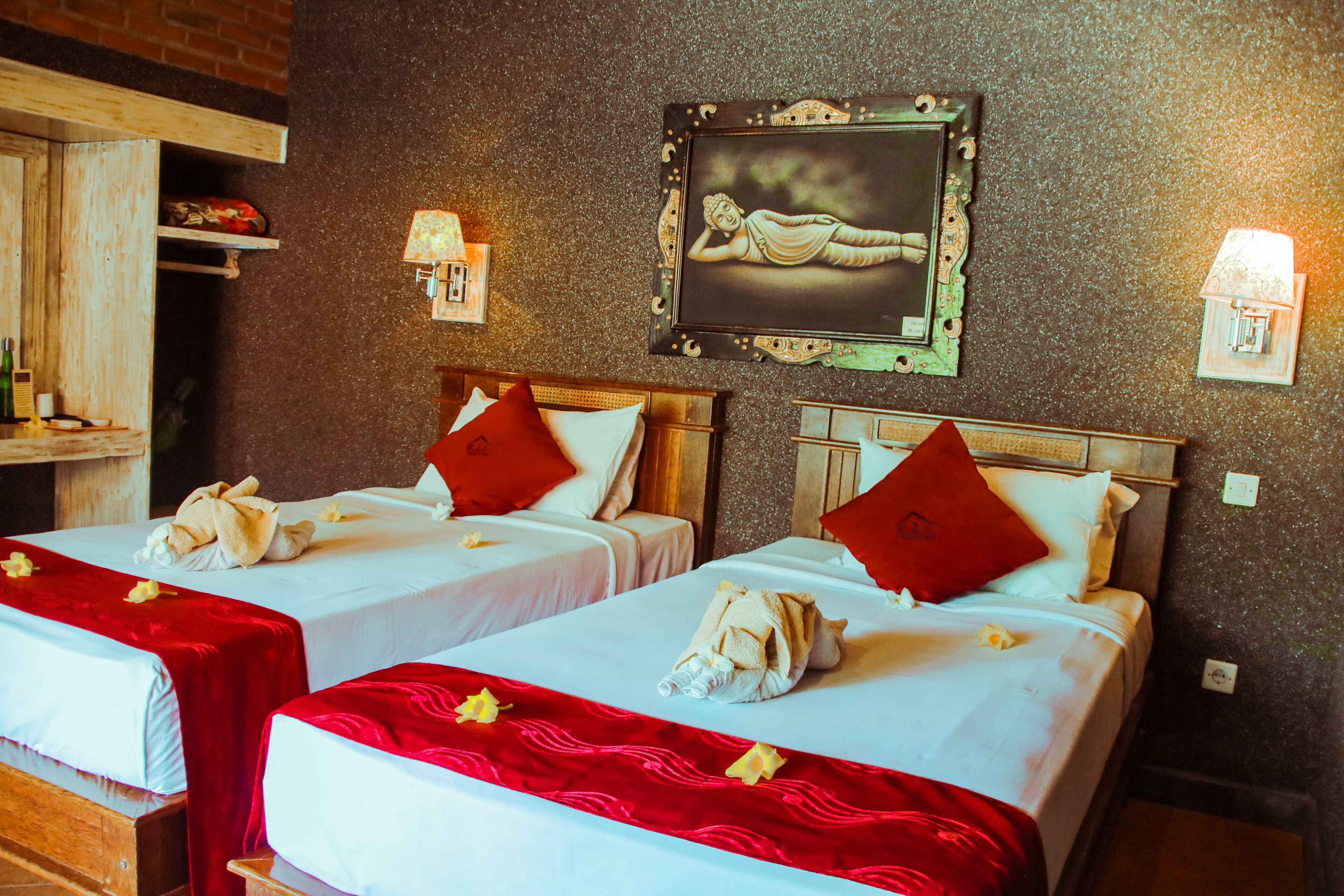 A hotel room with two beds, each with a red blanket and a white pillow. There is a framed picture on the wall and a small shelf with items on it.