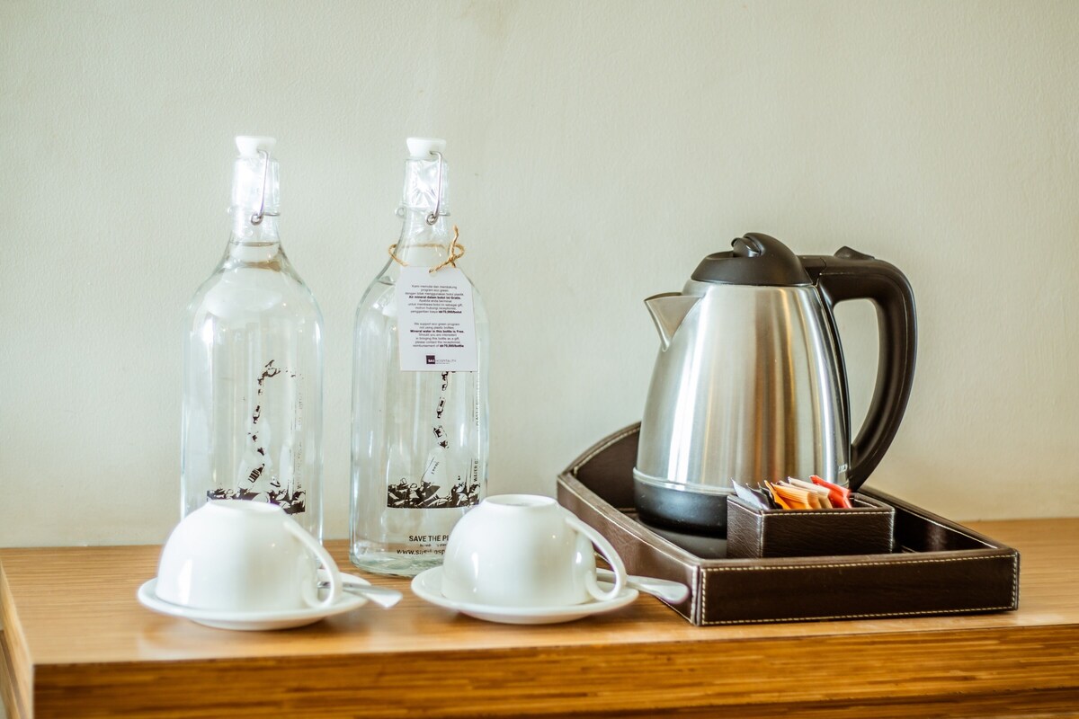 A wooden table with two clear bottles, a silver electric kettle, and two white cups with saucers.