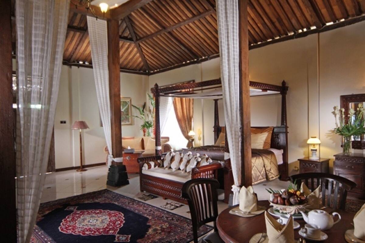 A traditional four-poster bed with a canopy, a sitting area with a sofa and armchairs, a dining table set for tea, and a large mirror on the wall.