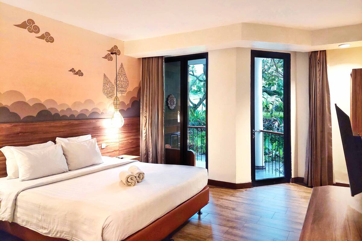 Une chambre d’hôtel avec un grand lit, une tête de lit en bois, une vue sur un balcon à travers une porte vitrée et une fresque murale décorative.