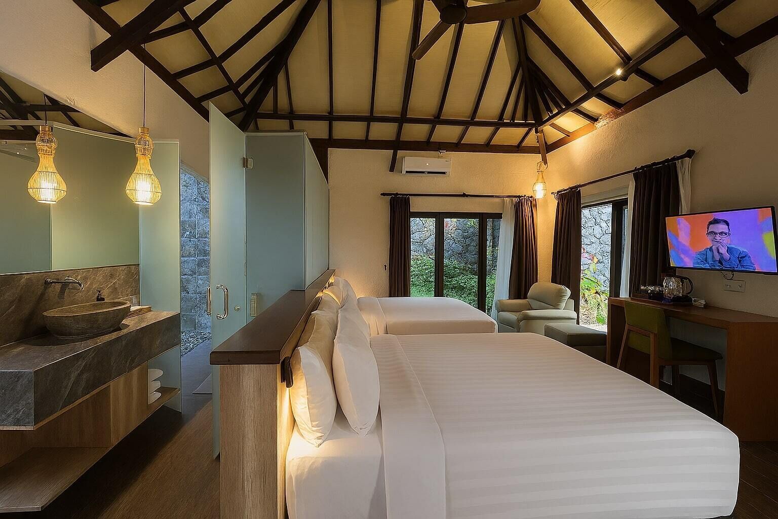 Habitación de hotel con cama, escritorio con televisor, baño con lavamanos y un amplio ventanal con cortinas.