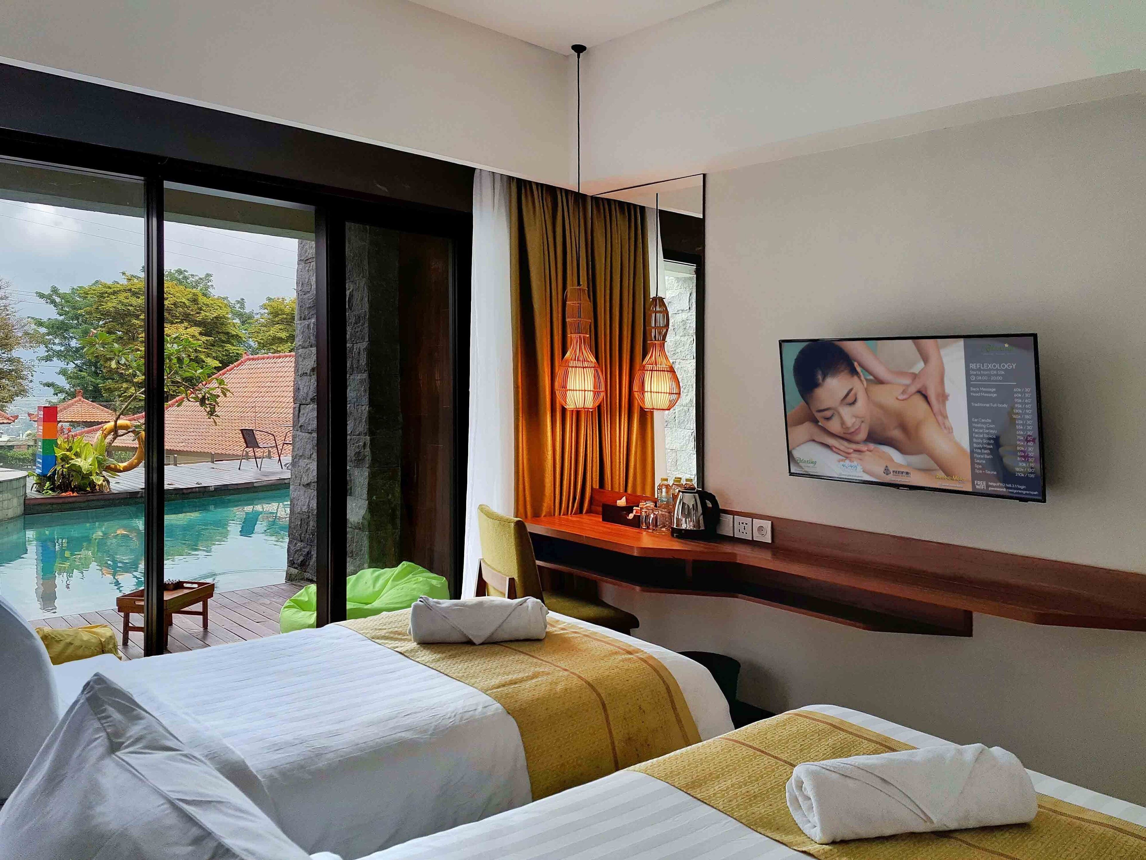 A hotel room with two beds, a TV displaying a reflexology service, a window with a view of a swimming pool and outdoor area, and a small table with a lamp and a bottle.