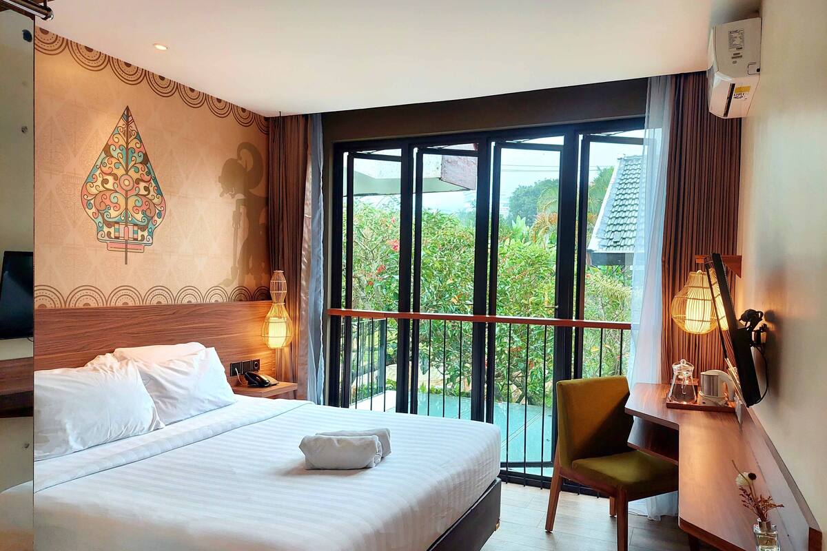 Une chambre d’hôtel avec un grand lit, un bureau avec une chaise, une télévision et un balcon donnant sur un espace verdoyant.