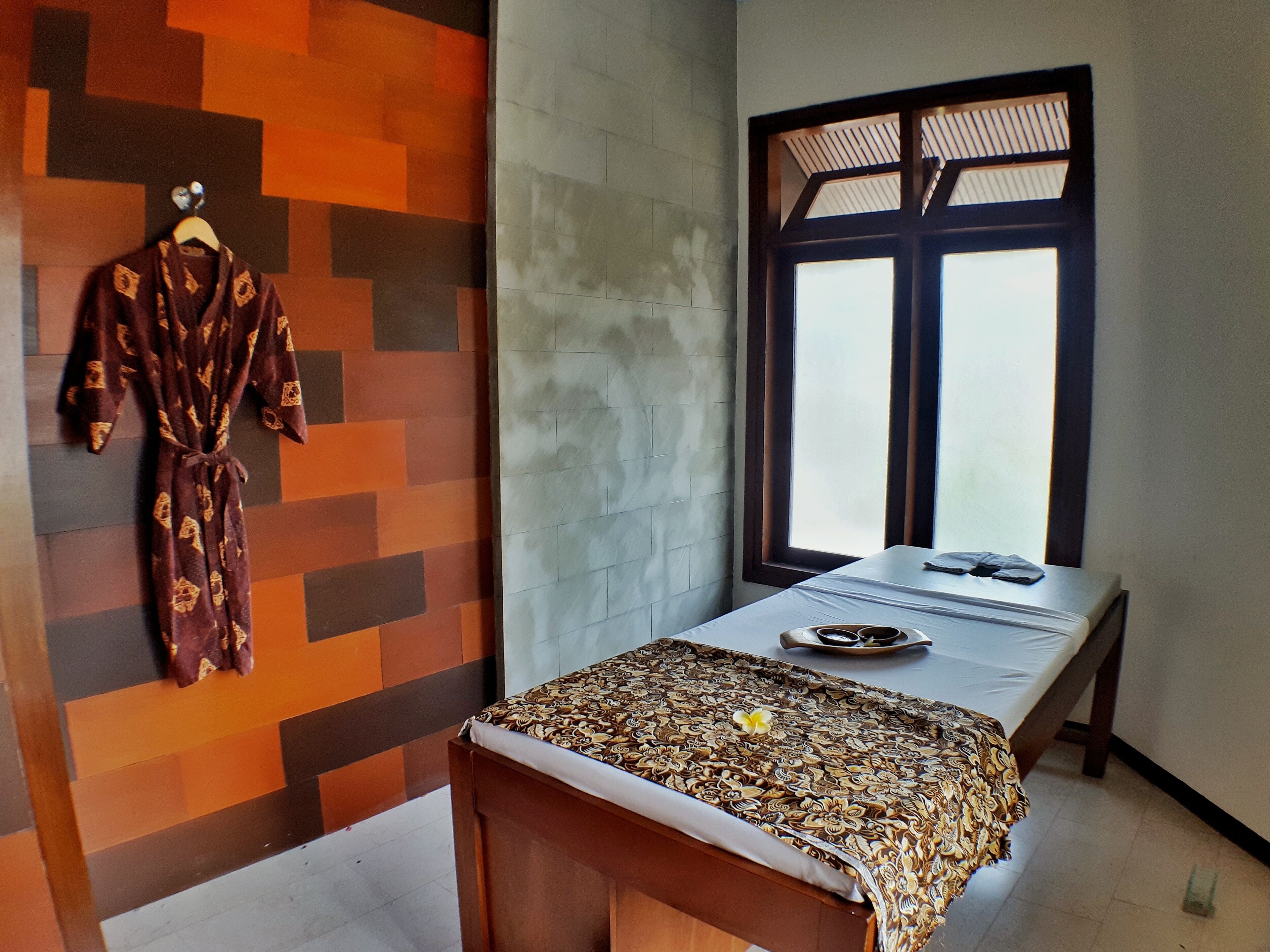 A room with a massage table, a patterned bedspread, a robe hanging on the wall, and a window with a view.