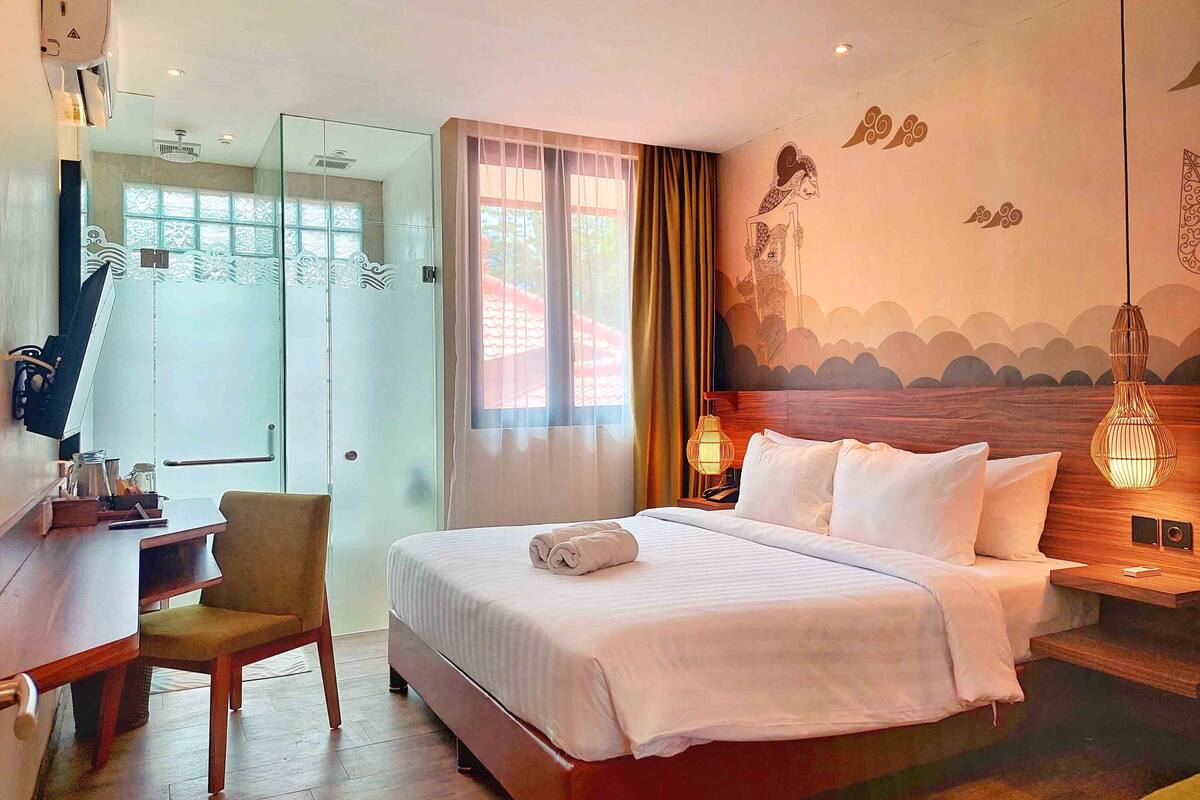 Une chambre d’hôtel comprenant un lit, un bureau, une chaise, une fenêtre avec des rideaux et un mur orné d’une fresque.