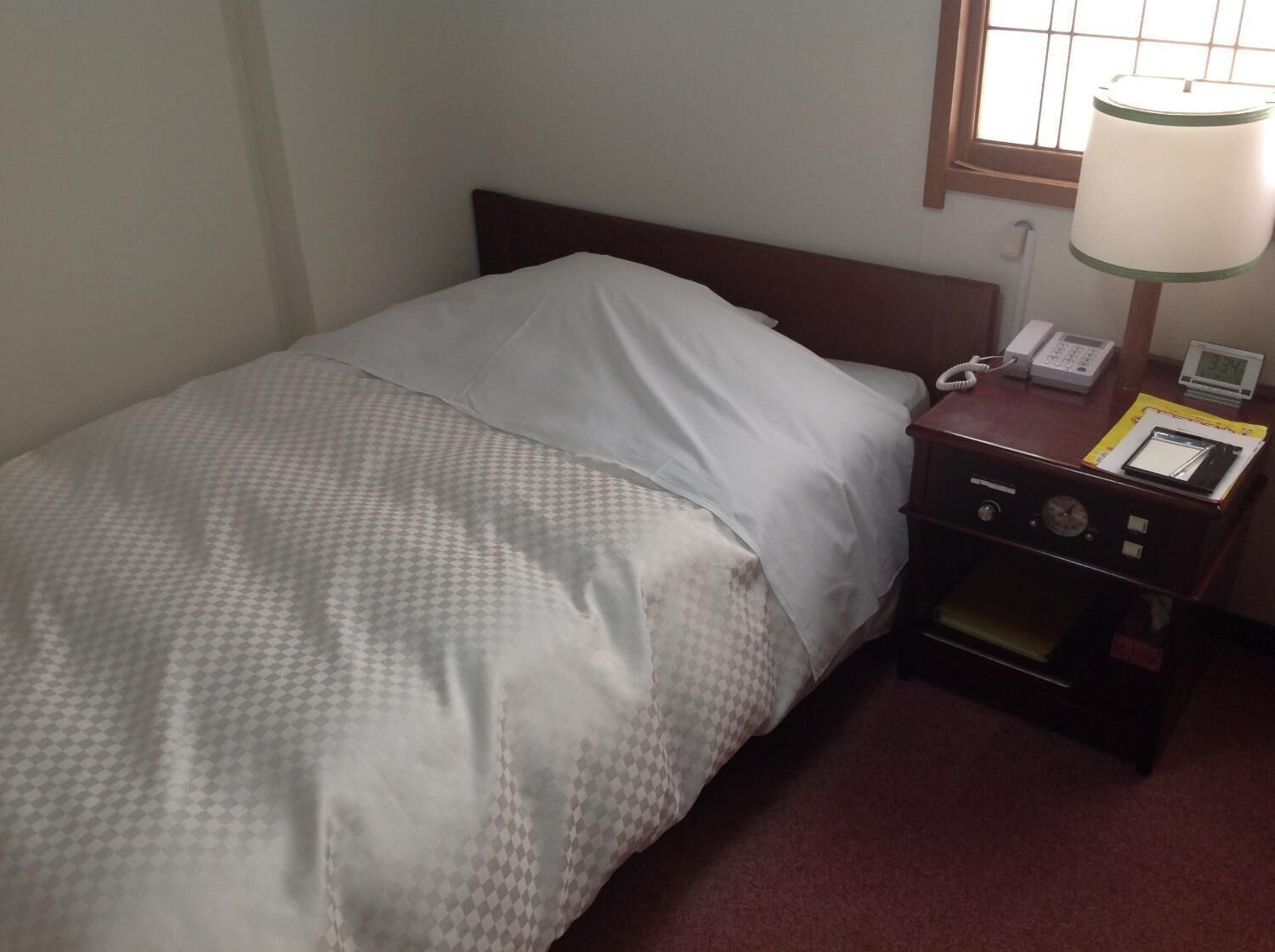 A hotel room with a single bed, a nightstand with a lamp, a phone, and a digital clock.